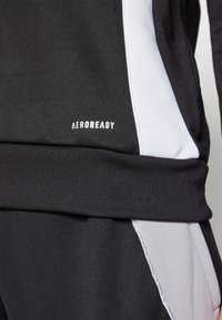 Fekete sportöltözet fehér oldalpanelekkel és texturált anyagból. "AEROREADY" logó fehér színben a derékvonal közelében.