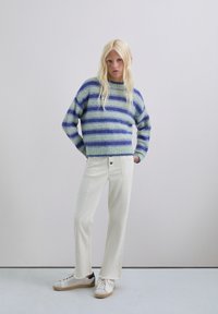 Suéter a rayas en verde y azul; textura suave. Combinado con unos vaqueros acampanados blancos y zapatillas blancas con acentos sutiles. Atuendo casual.