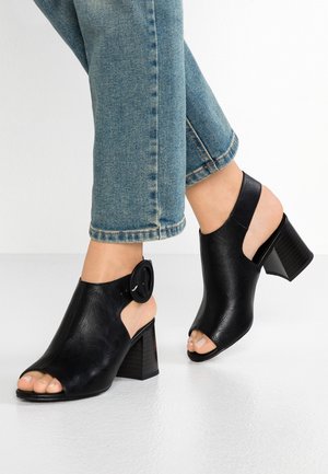 Pieds portant des chaussures noires à talon bloc ouvertes au niveau des orteils et du talon avec une grande boucle, associées à un jean bleu raccourci sur un sol blanc.