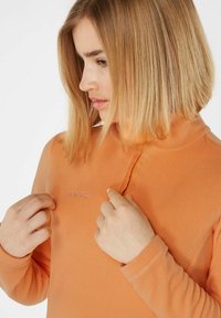 Pullover in polar orange con colletto alto e zip a metà, caratterizzato da un logo tono su tono sul petto e un morbido tessuto testurizzato.
