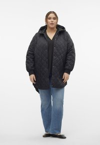 Chaqueta negra acolchada con capucha, que presenta un corte relajado, botones a presión y bolsillos laterales. Combinada con jeans acampanados azules y zapatos planos negros.