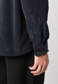 Donkerblauwe corduroy shirt met een ontspannen pasvorm, voorzien van een knoopmanchet en subtiele ribbelstructuur. Zichtbare hand die de zoom vasthoudt.