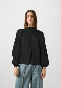 Blusa nera a maniche lunghe con collo alto, caratterizzata da dettagli in tessuto arricciato e polsini elastici, abbinata a pantaloni ampi blu chiaro.