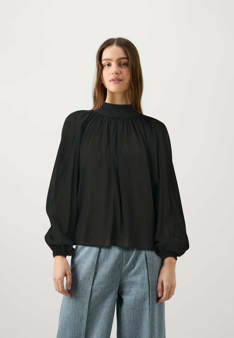 Blusa nera a maniche lunghe con collo alto, caratterizzata da dettagli in tessuto arricciato e polsini elastici, abbinata a pantaloni ampi blu chiaro.