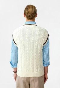 Crème-kleurige mouwloze trui met kabelpatroon, geribbelde zoom en contrasterende donkerbruine afwerking, gedragen over een blauw gestreept shirt.