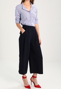 Modrá a bílá pruhovaná blůza s límečkem, kombinovaná s černými širokými kalhotami a červenými vysokými podpatky. Kalhoty mají boční kapsy.