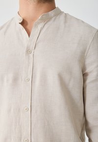 Lys beige linned skjorte med mandarinkrave, der har en knaplukning ned foran og en subtil tekstur.