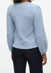 Light blue textured long-sleeve top with a round neckline and gathered sleeves, paired with dark denim jeans.