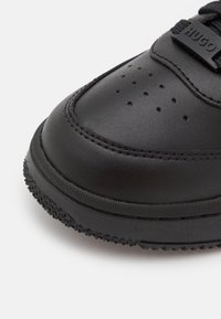 Bout de basket en cuir noir avec de petits trous de ventilation, coutures détaillées, semelle texturée et étiquette de lacet de marque indiquant "HUGO."