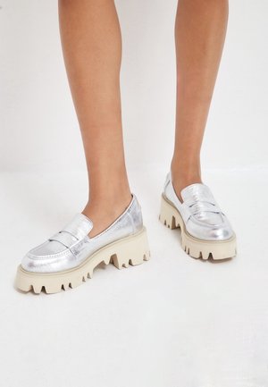 Pieds portant des mocassins métalliques argentés avec des semelles crantées écrues épaisses sur un sol blanc.