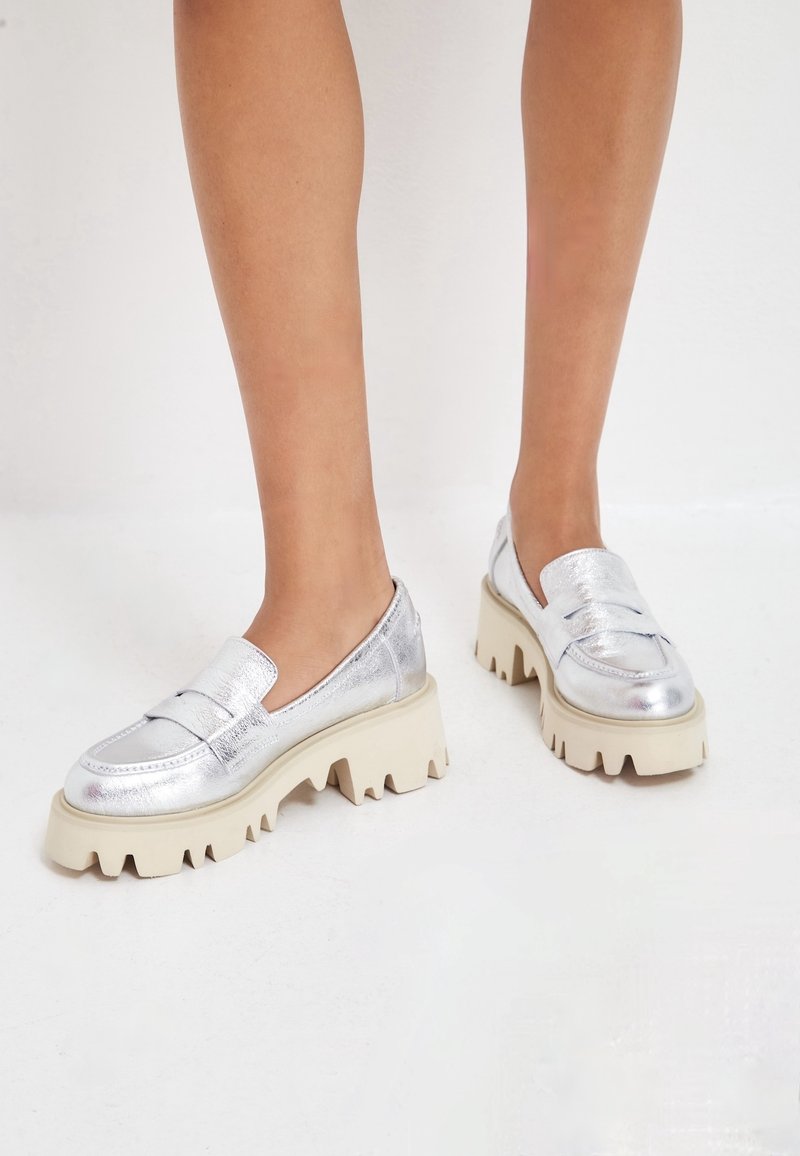 Pieds portant des mocassins métalliques argentés avec des semelles crantées écrues épaisses sur un sol blanc.