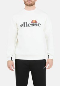 Sudadera blanca de mezcla de algodón, con un gran logo negro "ellesse" y un diseño en rojo y naranja arriba. Pantalones negros con un pequeño detalle del logo.