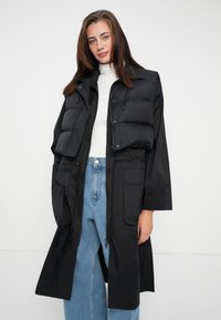 Lang zwarte donsjas met rits aan de voorkant, capuchon en meerdere zakken. Soepele textuur en oversized pasvorm, gelaagd over een witte top.