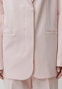 Femme portant un blazer cintré rose clair et un pantalon assorti avec des boutons cachés, montrant le bas du torse et les mains sur les côtés.