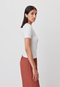 T-shirt bianca attillata a maniche corte con scollatura rotonda, abbinata a pantaloni plissettati color ruggine, presentata di profilo su uno sfondo neutro.