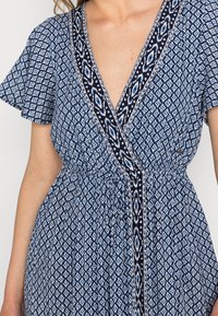 Blå mönstrad klänning med V-ringning, korta ärmar och dragsko i midjan. Tyget har ett diamantskärmönster med kontrasterande kant.