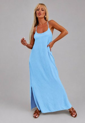 Femme aux longs cheveux blonds portant une robe longue bleu clair sans manches avec une fente latérale et des sandales marron sur un fond gris uni.