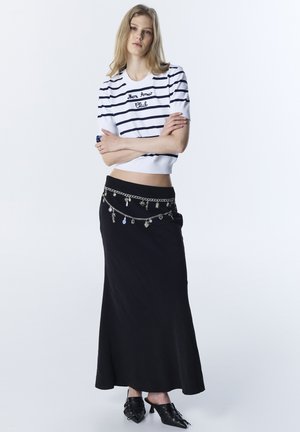 Mujer con los brazos cruzados, vestida con un top corto de rayas blancas y azul marino, falda larga negra con cinturón de cadenas decorativas en capas, y zapatos de tacón negros.