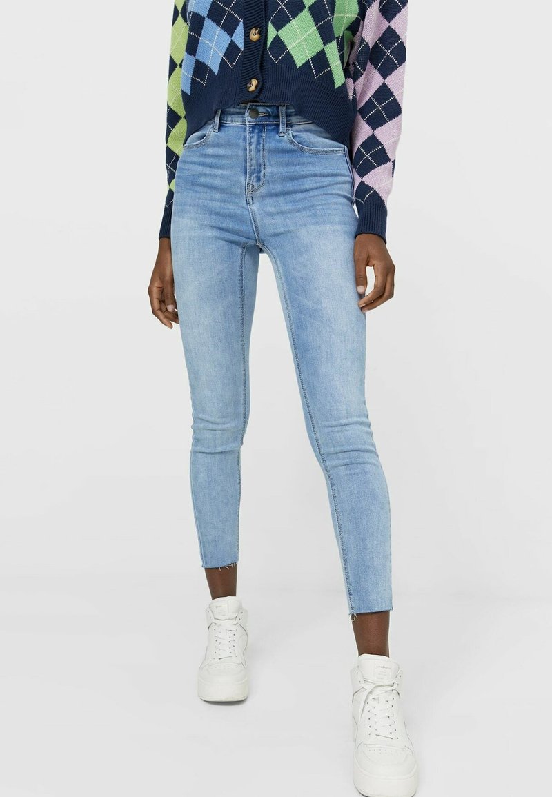 Jeans ajustados de color azul claro con cintura alta, con un dobladillo desflecado y diseño clásico de cinco bolsillos, combinados con zapatillas blancas.