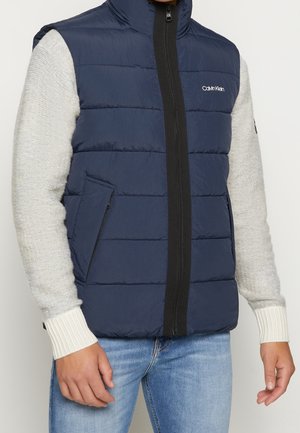 Homme portant une veste matelassée bleu marine Calvin Klein par-dessus un pull gris clair et un jean bleu, debout devant un fond uni.