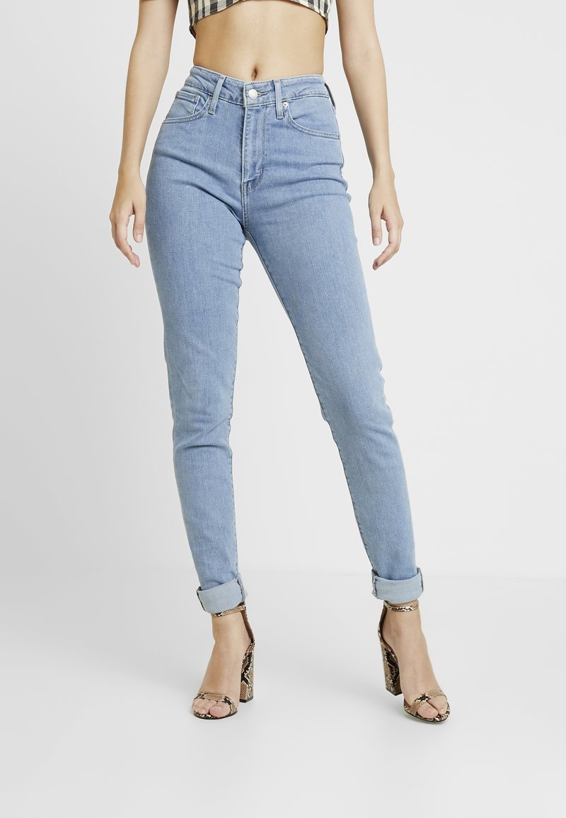 Jeans de tiro alto en azul claro con corte ajustado, cinco bolsillos y dobladillos remangados, combinados con sandalias de tacón con estampado de serpiente marrón.