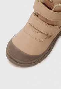 Botte beige pour enfants avec un cuir marron à l'avant et deux sangles Velcro ajustables sur fond blanc.