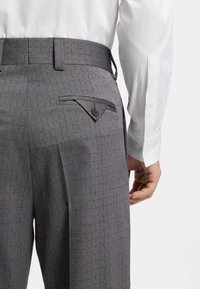 Grijze textuurachtige broek met een patroonweefsel, voorzien van een decoratieve achterzakklep en een knoopsluiting. Een wit overhemd is gedeeltelijk zichtbaar.