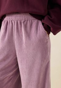 Pantalons en velours côtelé violet clair avec une ceinture smockée, présentant des côtes verticales et des poches latérales, portés avec un haut bordeaux foncé.