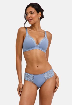 Frau posiert in hellblauem Lingerie-Set mit Spitzen-Details an den BH-Cups und dem Slip vor schlichtem hellem Hintergrund.