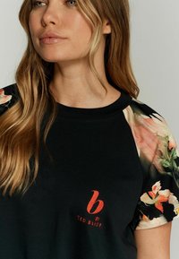 Melna t-kreklu ar ziedu raksta piedurknēm. Uz krūtīm ir sarkans izšuvums "b by TED BAKER". Mīksta auduma, brīvi pieguļoša.
