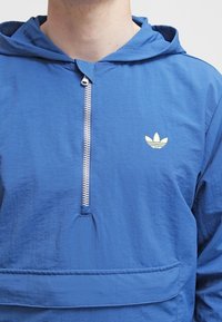 Modrá lehká bunda s kapucí a půlzipem vpředu, vybavená přední kapsou a malým žlutým logem na levé straně hrudi.