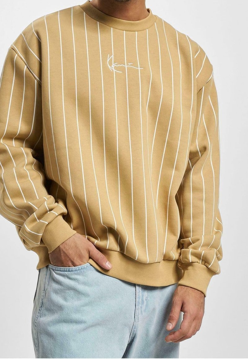 Homme portant un sweat-shirt beige à col rond avec des fines rayures blanches verticales et un jean bleu clair, une main dans la poche.