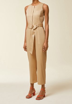 Combinaison beige sans manches avec poche avant et ceinture nouée, portée avec des sandales à talons blocs orange à brides, sur une personne debout.