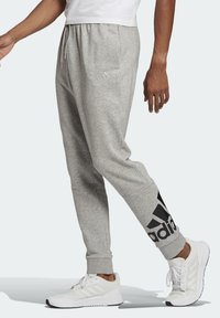 Mand iført grå Adidas sweatpants med sort logo på læggen, hvide sneakers og en hvid skjorte, der står sidelæns på en lys baggrund.