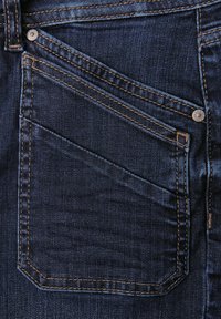 Jeans de mezclilla oscura con un gran bolsillo trasero con costuras diagonales, costuras de cobre y un acabado de botón de metal. Textura suave y líneas limpias.