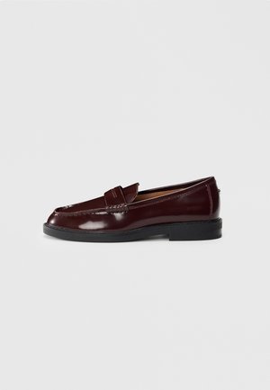 Mocassin penny loafer en cuir marron foncé avec semelle noire, talon bas et détail de couture le long du bord supérieur, présenté en profil latéral.