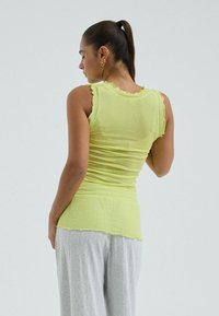 Mujer con cabello oscuro recogido en una coleta, vestida con una camiseta sin mangas de color amarillo claro y pantalones anchos grises, vista de espaldas sobre un fondo liso.