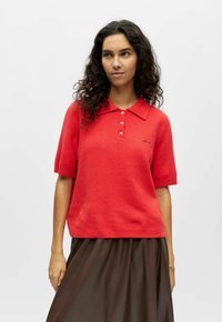 Pull polo rouge  à manches courtes avec col, trois boutons et finition texturée. Associé à une jupe marron. Détail subtil du logo sur la poitrine.