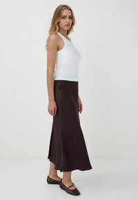 Un haut sans manches blanc associé à une longue jupe brune, présentant une texture lisse et une fente sur le côté. Des ballerines noires tressées complètent la tenue.