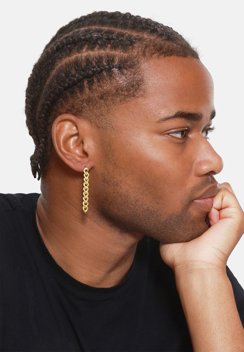 Gold chain-link earring hangs from a lobe, featuring a textured design. The individual has braided hair and is resting his chin on his hand.