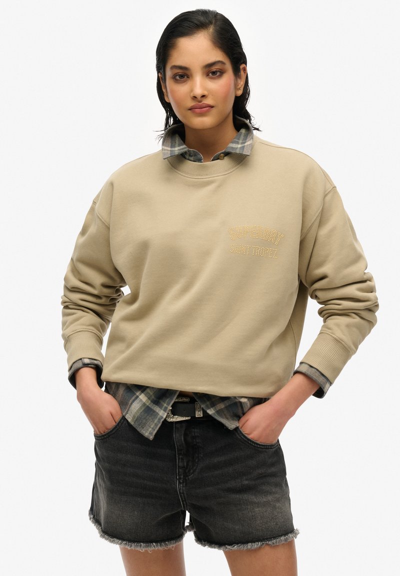 Sudadera beige con puños acanalados y el logo "Superdry Saint Tropez". Llevada sobre una camisa de cuadros en capas y combinada con pantalones cortos de mezclilla oscuros.