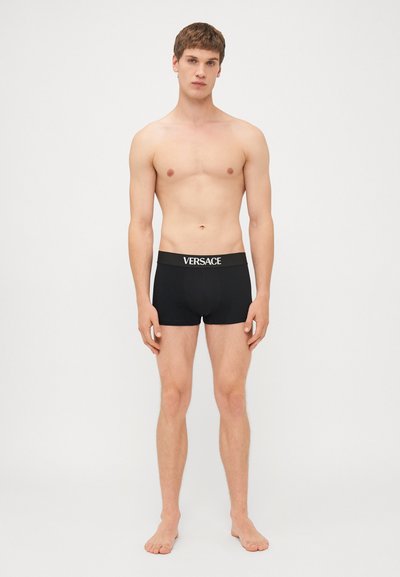 Sorte boxer shorts med en bred sort elastikliv, der har "VERSACE" skrevet i hvidt, lavet af et blødt, glat stof og med en tætsiddende design.