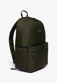 Donkergroene rugzak met voorvak met ritssluiting, zwarte onderkant, gevoerde schouderbanden en "Superdry Outdoor Originals" logo patch aan de voorkant.