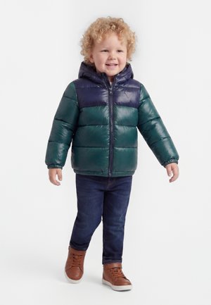 Toddler with curly blond hair wearing a green and navy puffer jacket, dark jeans, and brown shoes, smiling and walking forward.