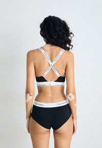 Svart Calvin Klein bralette og høytlivs truse med hvite logobånd, med kryssryggdesign og glatt tekstur.