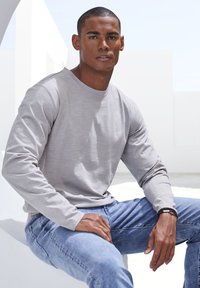 Helles graues Langarmshirt mit Rundhalsausschnitt, kombiniert mit blauen Jeans. Glatte Stofftextur, minimalistisches Design, sitzt vor einem weißen Hintergrund.