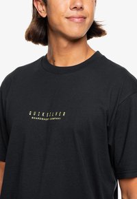 Mustat lyhythihaiset t-paidat puuvillasta, jossa on keltainen "Quiksilver" -logo ja "Boardshort Company" -teksti modernilla fontilla.
