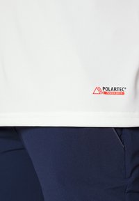 Faldón de prenda blanca con el logo rojo de Polartec Power Dry, llevado sobre pantalones azul marino con un bolsillo lateral visible y una costura.