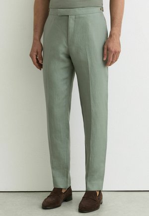 Jasnozielone, dopasowane spodnie z bocznymi kieszeniami, noszone z ciemnobrązowymi zamszowymi mokasynami, przedstawione na stojącym męskim modelu na białym tle.