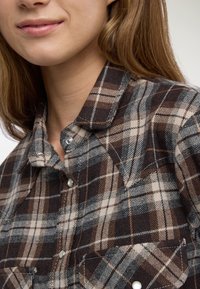 Giovane donna che indossa una camicia a quadri marrone, beige e nera con bottoni a pressione e colletto, mostrata dal mento al petto.